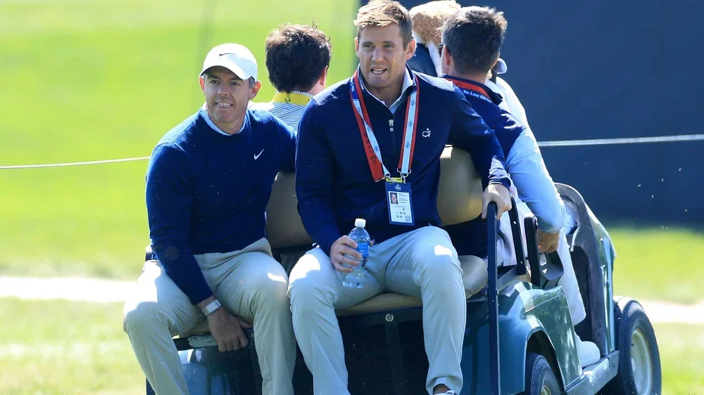 Four men in golf attire and event lanyards ride together on a golf cart near a green field at the PGA Championship, with one holding a water bottle and another sitting on the edge, all looking in different directions.